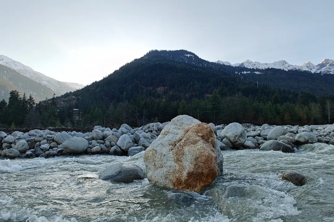 A calm river with scattered rocks in Solang Valley, reflecting the theme of "A Reminder Carved in Stone" by Sadhuwani